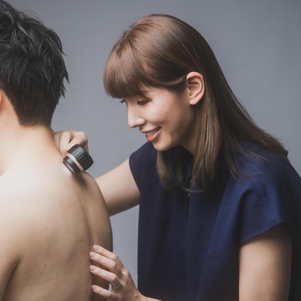 A woman uses a handheld device on a man's back while smiling, promoting awareness for skin cancer prevention. The man has his back turned towards the camera.