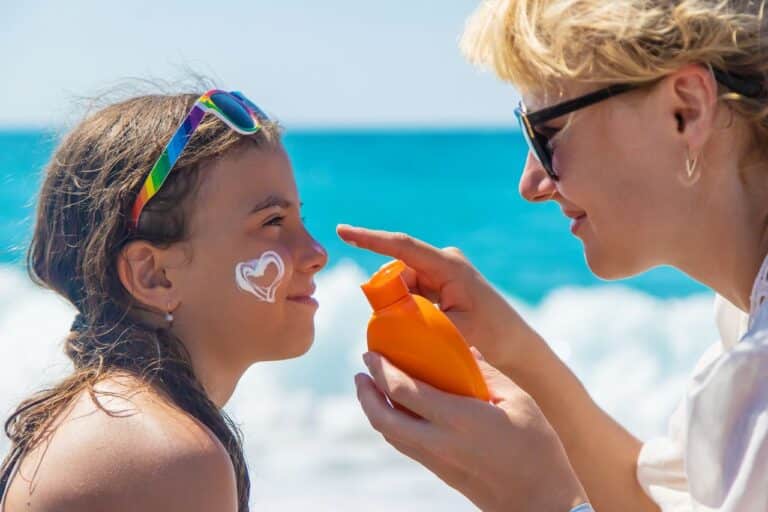 A woman applies sunscreen to a smiling girl on her cheek at the beach emphasising preventive treatments for conditions like solar keratosis.