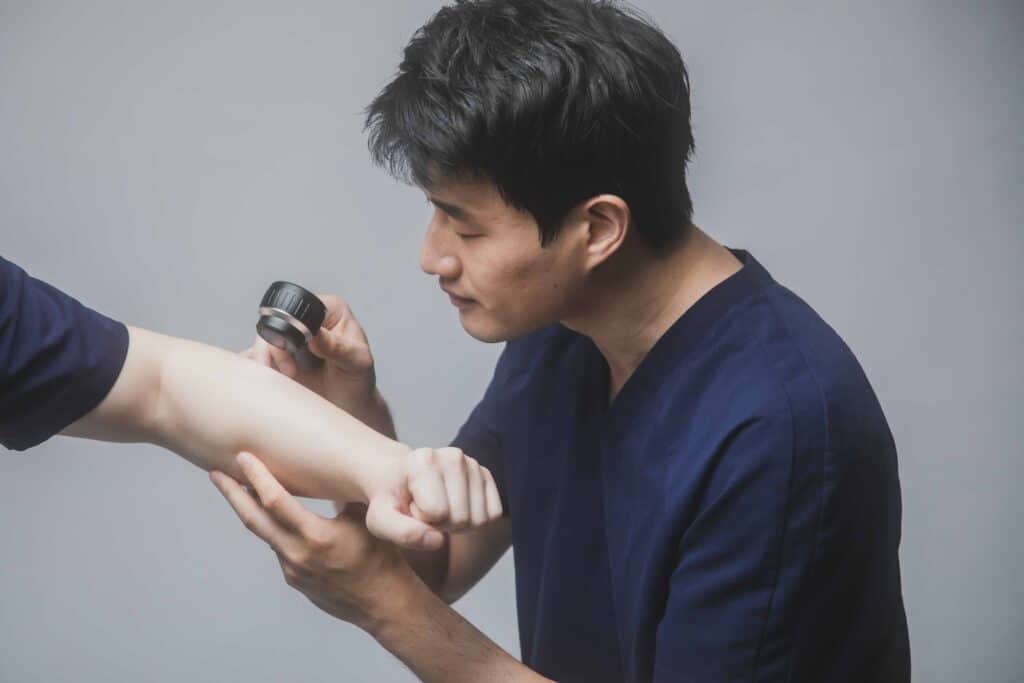 A medical professional examines a person's arm using a magnifying tool, likely for a skin condition, against a plain background.