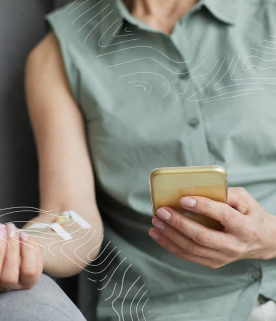 Person with an IV Infusion for enhanced nutrients on their arm holds a smartphone while sitting.