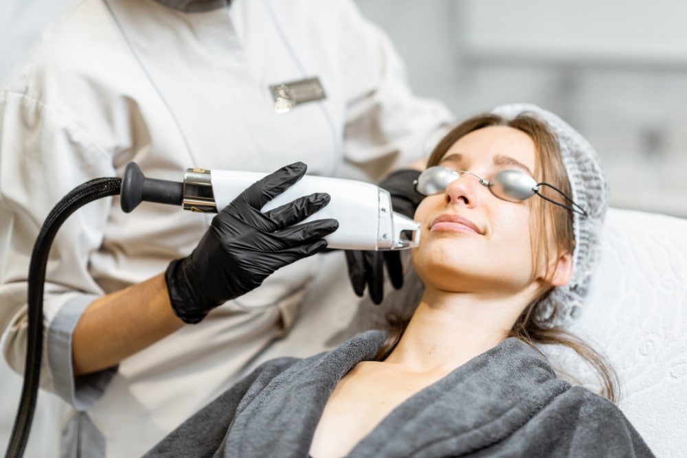 A woman receiving a non-ablative laser skin resurfacing treatment on her face at a clinic. She is wearing protective eyewear and a gray robe, while the practitioner, dressed in a uniform and black gloves, carefully administers the procedure.