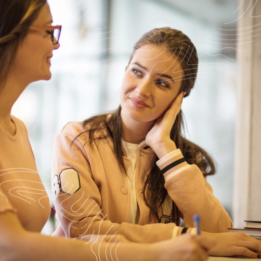 Two women, both medical professionals, sit at a table—one writing and the other smiling while listening. Both are casually dressed and engaged in conversation.