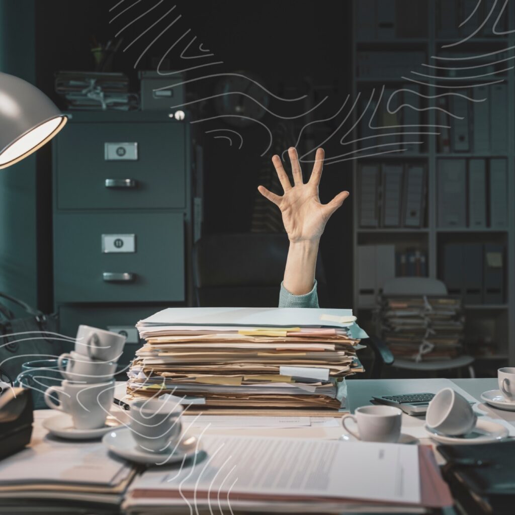 A hand reaching up from behind a large stack of paperwork on a cluttered desk, with coffee cups, a desk lamp, and filing cabinets in the background—a typical scene for busy medical professionals.