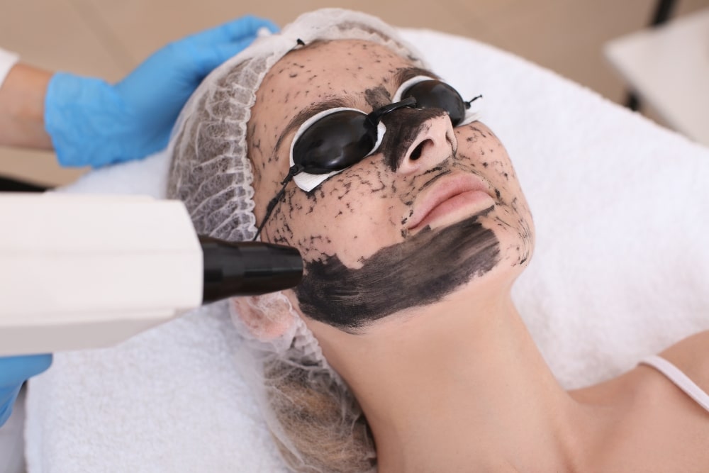 A person receives a facial laser treatment service while wearing protective eyewear and a hair cover. The face is partially covered with a dark treatment mask.