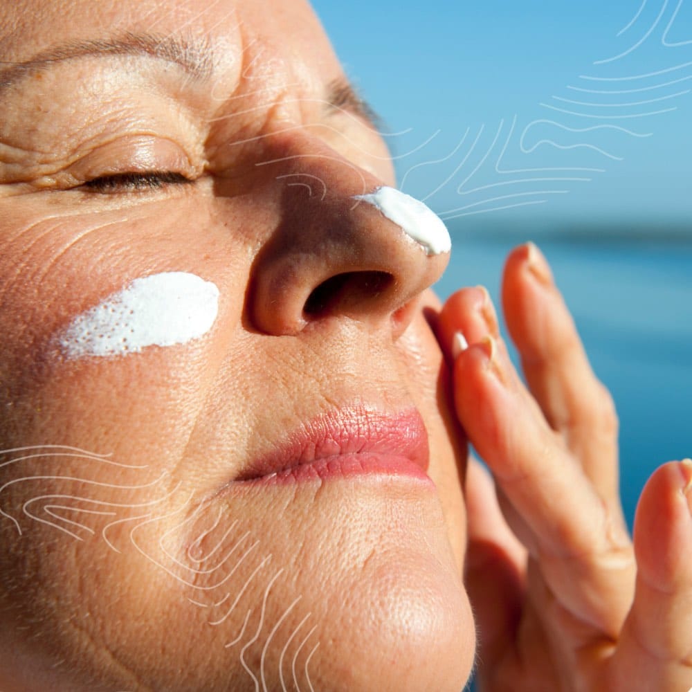 Close-up of a person applying sunscreen to their face, including their nose and cheeks, with eyes closed against a bright blue background.