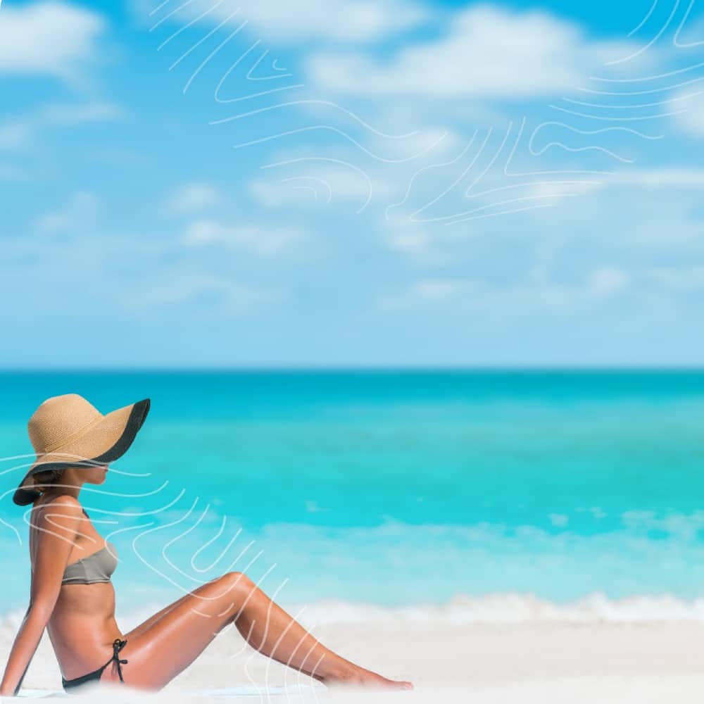 A person in a bikini sits on a sandy beach with a turquoise ocean and bright blue sky in the background, wearing a wide-brimmed hat. They seem at home in this serene paradise.