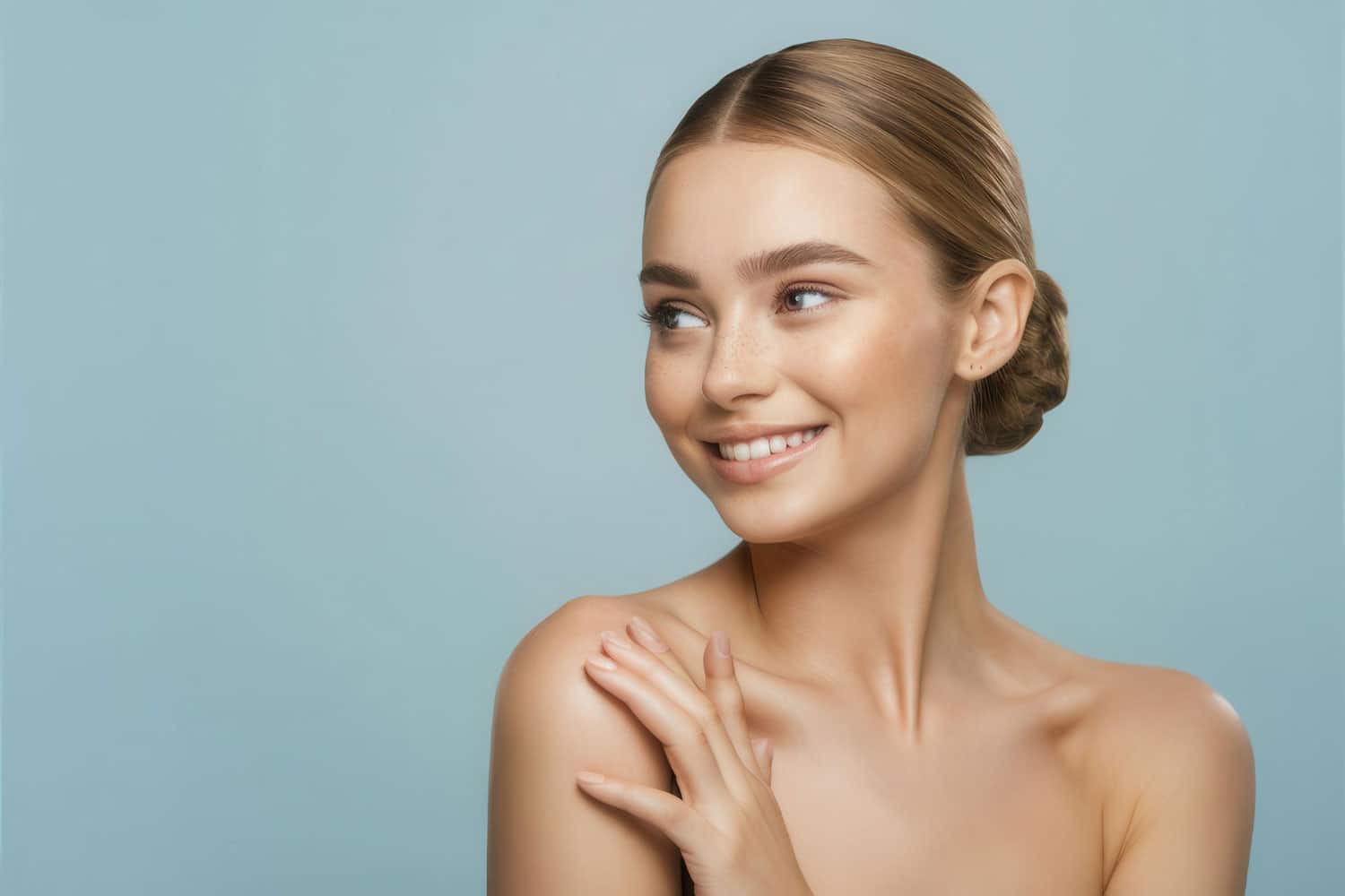 Young woman with a smile, light skin, and sleek hair in a bun, posing against a light blue background. She has one hand resting on her shoulder and is looking off to the side, radiating confidence possibly due to recent scarless mole removal.