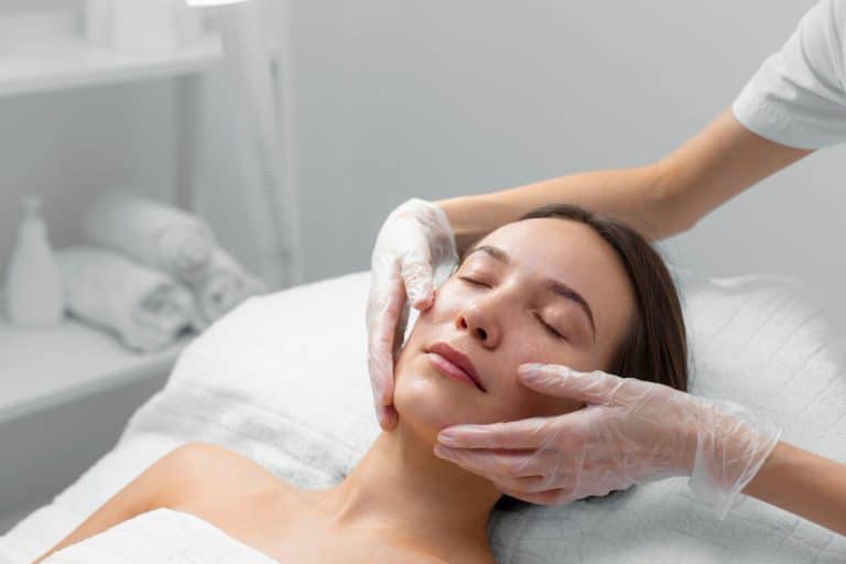 A serene scene unfolds as someone enjoys a soothing facial massage on a spa bed, gloved hands gently tending to their skin. In the tranquil backdrop, towels are neatly stacked, creating an inviting atmosphere where everyone feels pampered and rejuvenated.