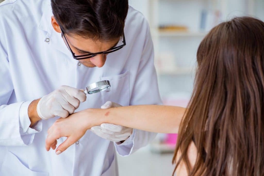 A doctor wearing gloves examines a patient's arm with a magnifying glass, focusing on a visible skin mark or rash that may indicate risk of skin cancer.