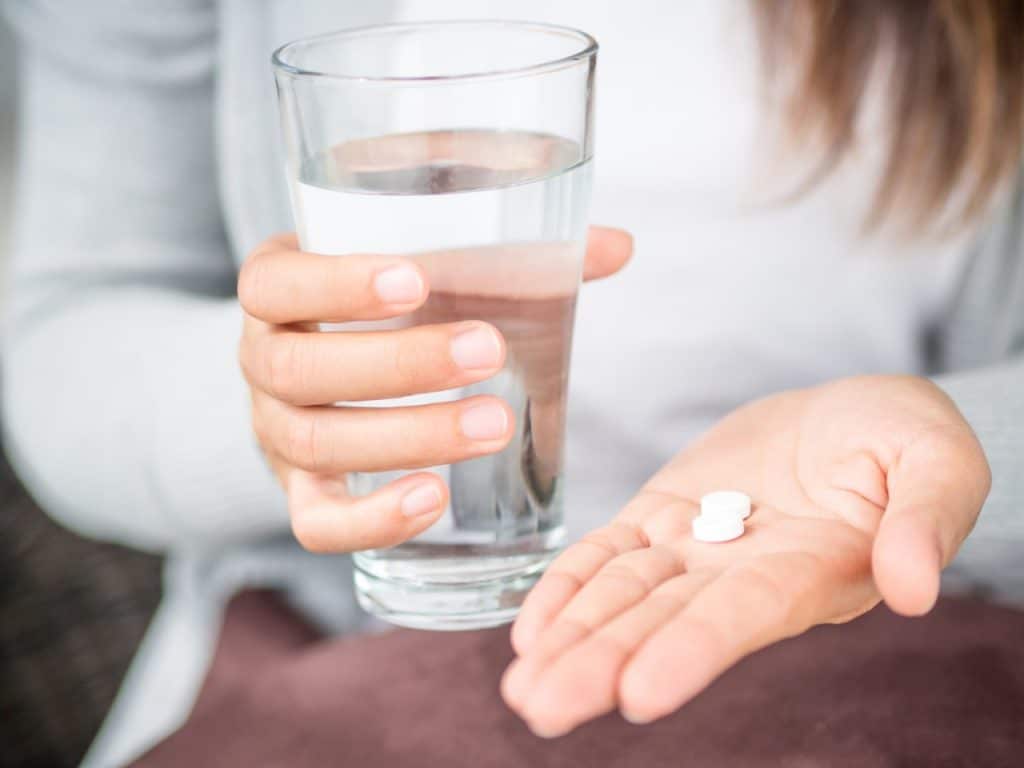 A person holds two white tablets in one hand and a glass of water in the other, considering treatment options for symptoms of Melanoma.