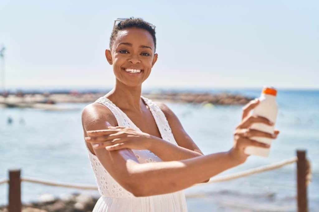A woman in a white dress stands outdoors by the water, smiling while applying sunscreen to her arm, protecting her skin on a sunny day.