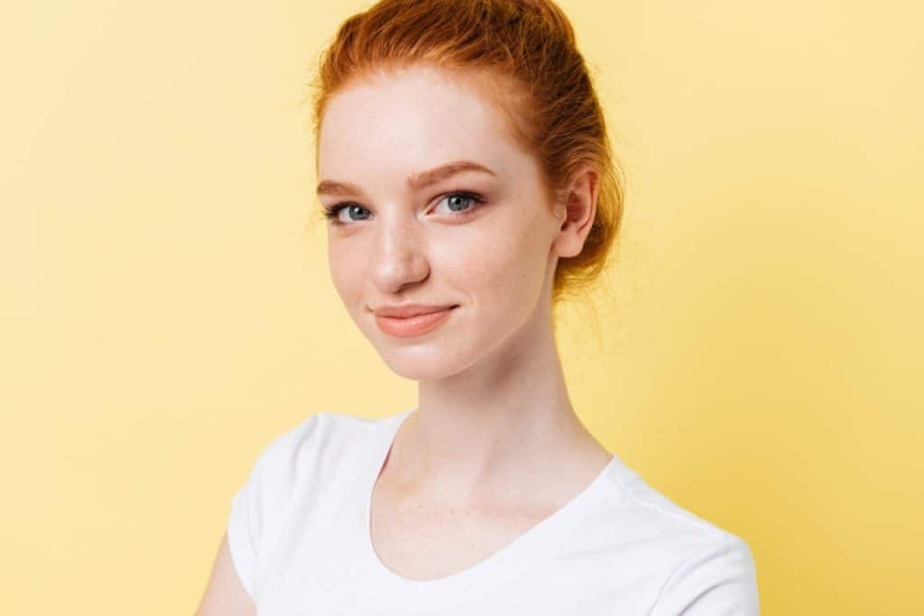 Young woman with red hair and fair skin, wearing a white t-shirt, smiling slightly, stands in front of a plain yellow background—features often highlighted in discussions about non-melanoma skin cancers like basal cell carcinoma.