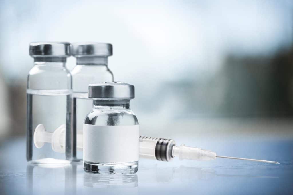 Three vaccine vials and a filled syringe, similar to those used for tanning injection treatments like Melanotan, are placed on a reflective surface with a blurred background.
