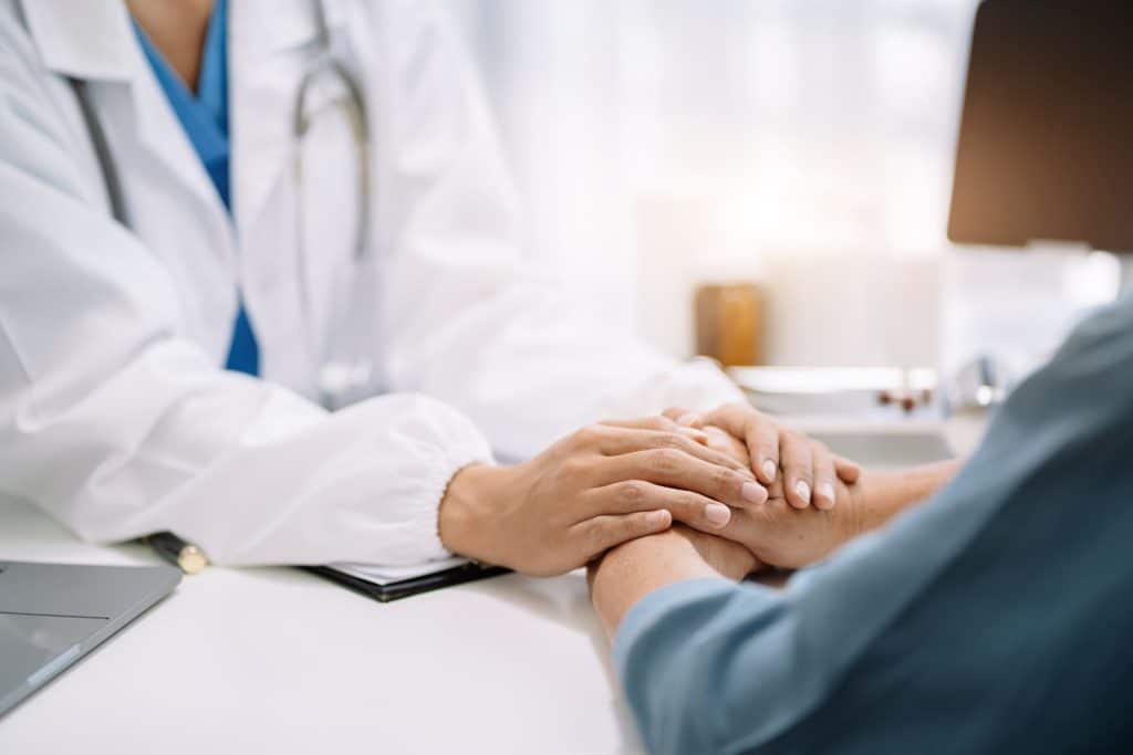 A doctor in a white coat holds a patient's hands aware of psychological impact in discussing the diagnosis and potential skin cancer risk factors.