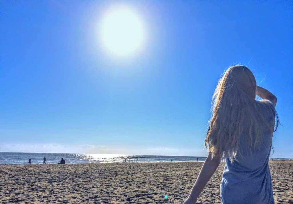 Person with long blonde hair stands on a sandy beach under a bright sun, looking toward the ocean—a scene shaped by coastal geography where high UV exposure can impact skin cancer rates. A few people are visible in the distance near the shoreline.