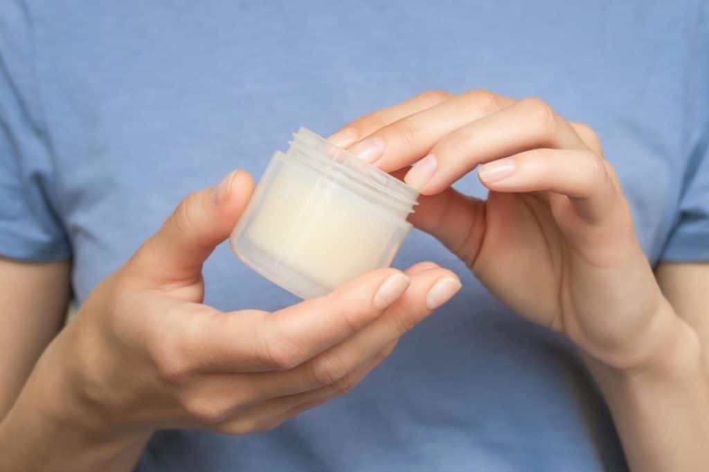 A person in a blue shirt holds an open jar of cream or ointment, with one hand dipping a finger into the product—a practical guide to wound dressings and post-surgical wound care.