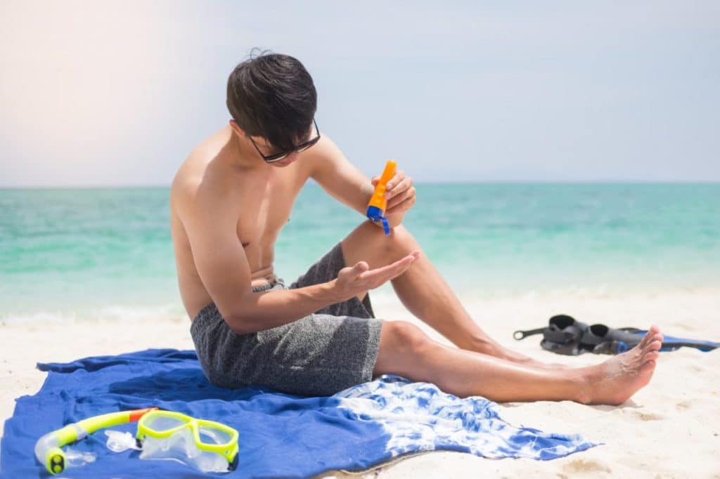 A man sits on a blue towel at the beach, applying sunscreen to his arm. He is wearing sunglasses and swim trunks, with snorkeling gear and fins beside him—busting the myth that Asians don’t need protection from skin cancer.