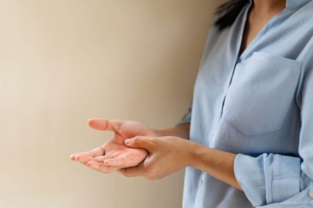 A person in a light blue shirt holds their right wrist with their left hand, standing against a plain beige background—challenging the myth that Asians aren’t at risk for skin cancer.