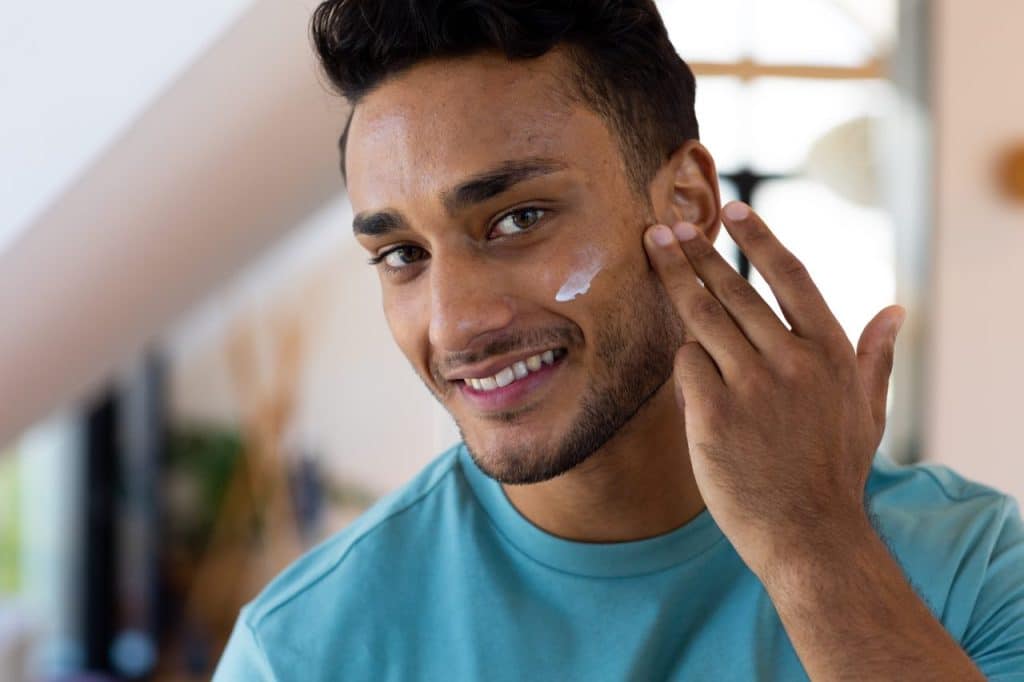 A tanned skin man in a turquoise shirt is smiling while applying lotion to his cheek indoors, taking steps toward skin cancer prevention for all skin types.