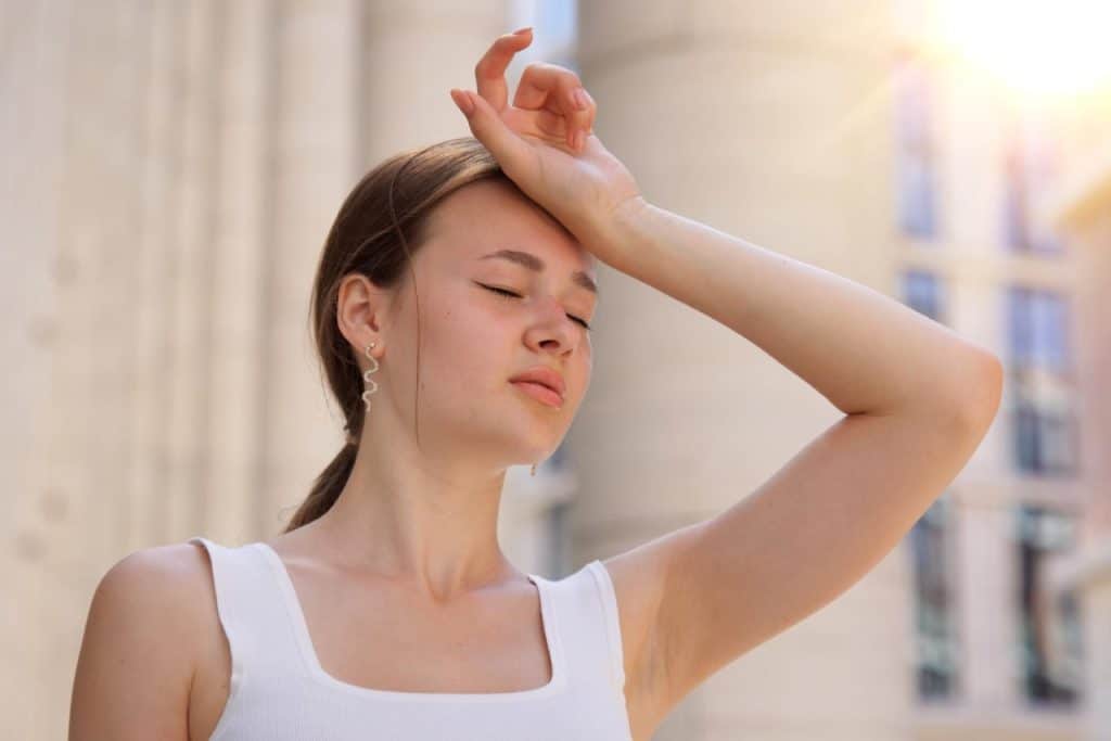 A woman in a white tank top stands outside with her eyes closed and hand on her forehead, appearing to feel hot or fatigued under bright sunlight, possibly due to strong UVB rays that can increase the risk of skin cancer.