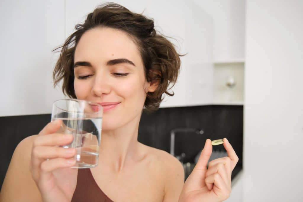 A woman holds a glass of water in one hand and a supplement pill for Vitamin D deficiency in the other, standing in a modern kitchen.