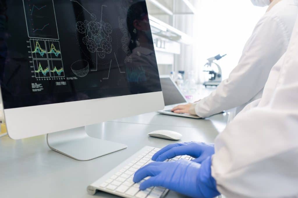 Scientist wearing blue gloves types on a keyboard in a laboratory, analysing AI data and molecular diagrams on a large monitor to drive breakthroughs in skin cancer treatment. 