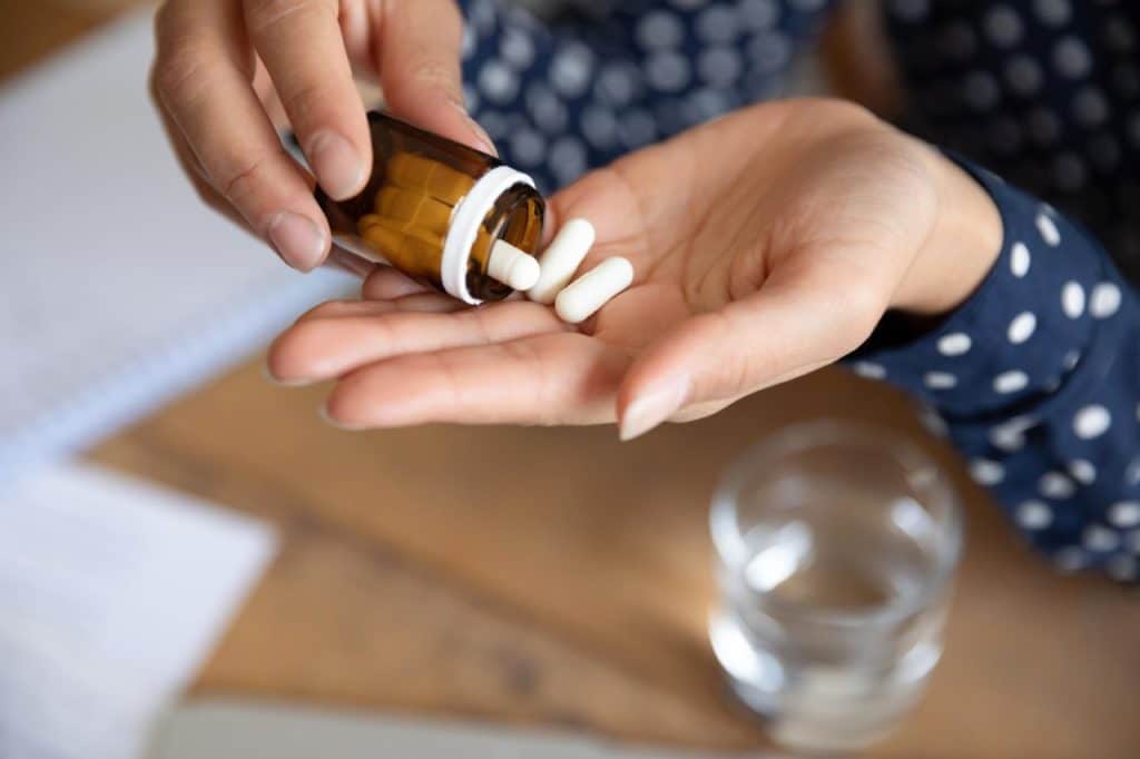 A person pours white capsules, used to manage immunosuppression, from a brown pill bottle into their hand, with a glass of water and a notebook on the table.