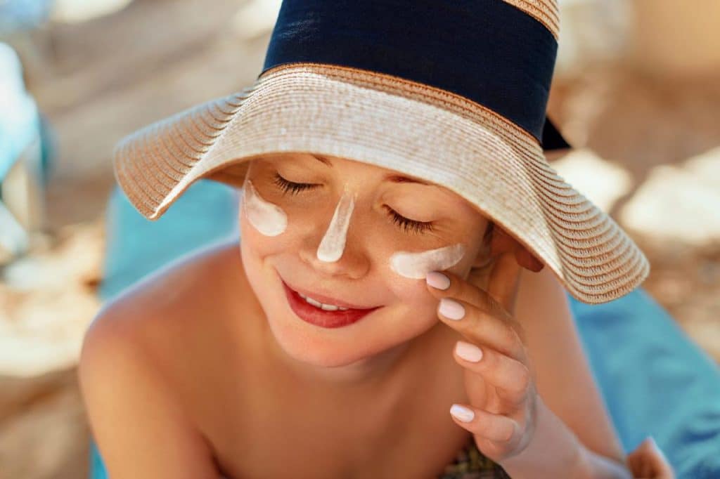 A person wearing a wide-brimmed hat applies sunscreen to their face while sitting outdoors, eyes closed and a slight smile, protecting their skin from ultraviolet light.