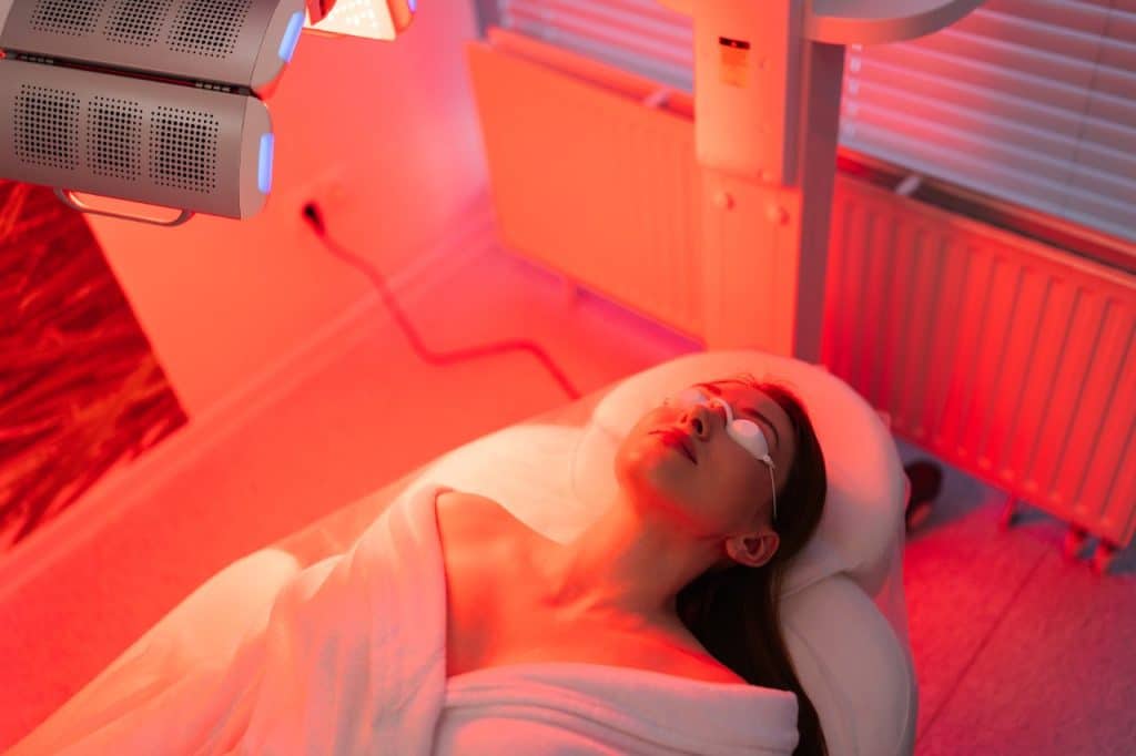 A woman lies on a treatment bed under red LED lights, wearing protective goggles and wrapped in a towel, experiencing the latest innovations in a clinical or spa setting.