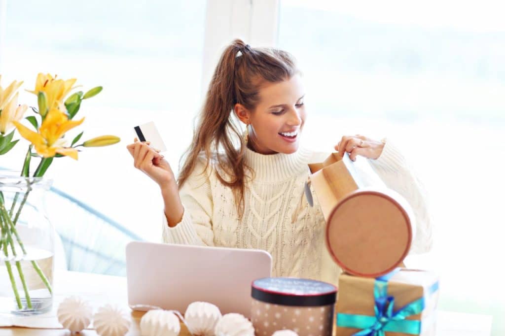 A woman sits at a table with a laptop, holding a credit card and smiling while opening a gift box during the June Campaign. Wrapped gifts, flowers, and a Spin the Wheel sign hint that Everyone’s a Winner this season.