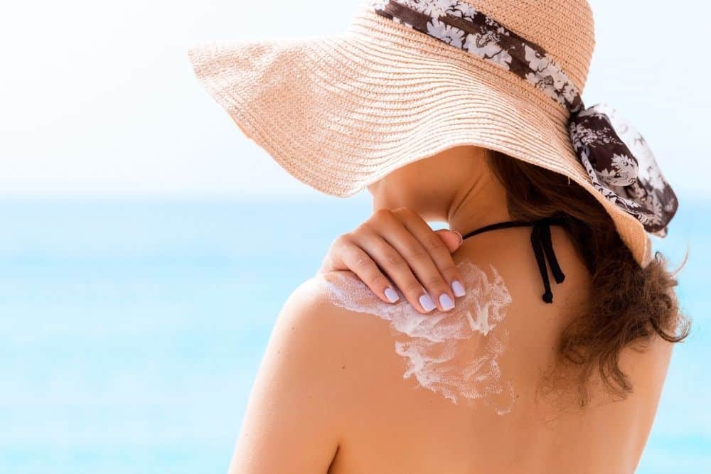A woman in a sun hat applies sunscreen to her shoulder while standing by the ocean, practicing skin cancer prevention.