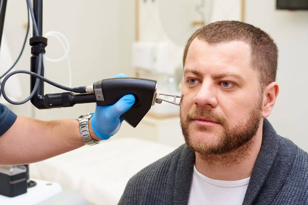 A man undergoes an examination with a specialised medical device, similar to those used in PDT or Fraxel Laser treatments, operated by a healthcare professional wearing blue gloves.