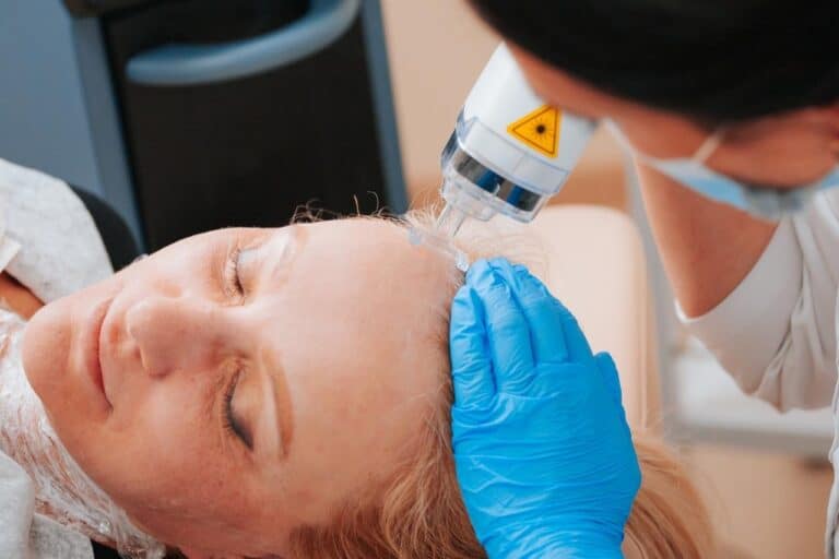 A woman lies with her eyes closed while a medical professional in blue gloves performs a Fraxel laser cosmetic procedure on her forehead to treat Basal Cell Carcinoma.