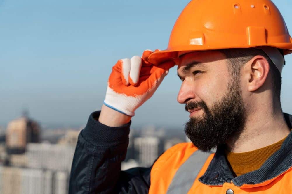 A construction worker wearing an orange safety helmet and vest adjusts his helmet while standing outdoors with buildings in the background, highlighting the importance of Skin Cancer Prevention under intense sun exposure.