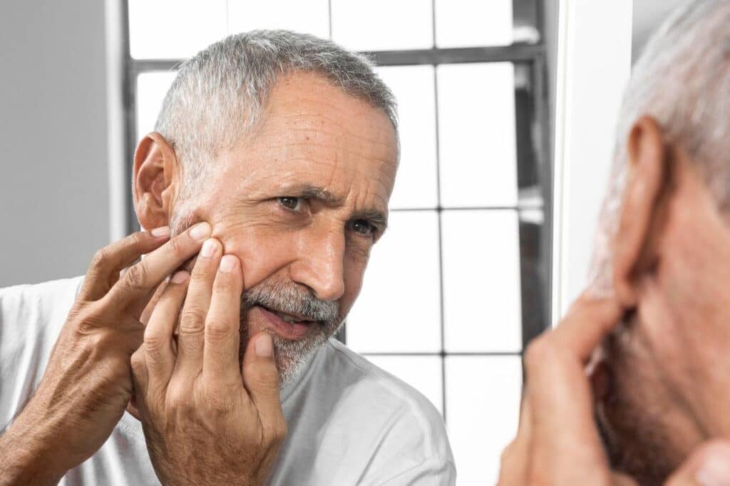 An older man with gray hair examines his cheek closely in a bathroom mirror, touching his face with his fingers as he inspects for lack-of-collagen-causing-dry-skin and increased risk of photo damaged skin leading to skin cancer.