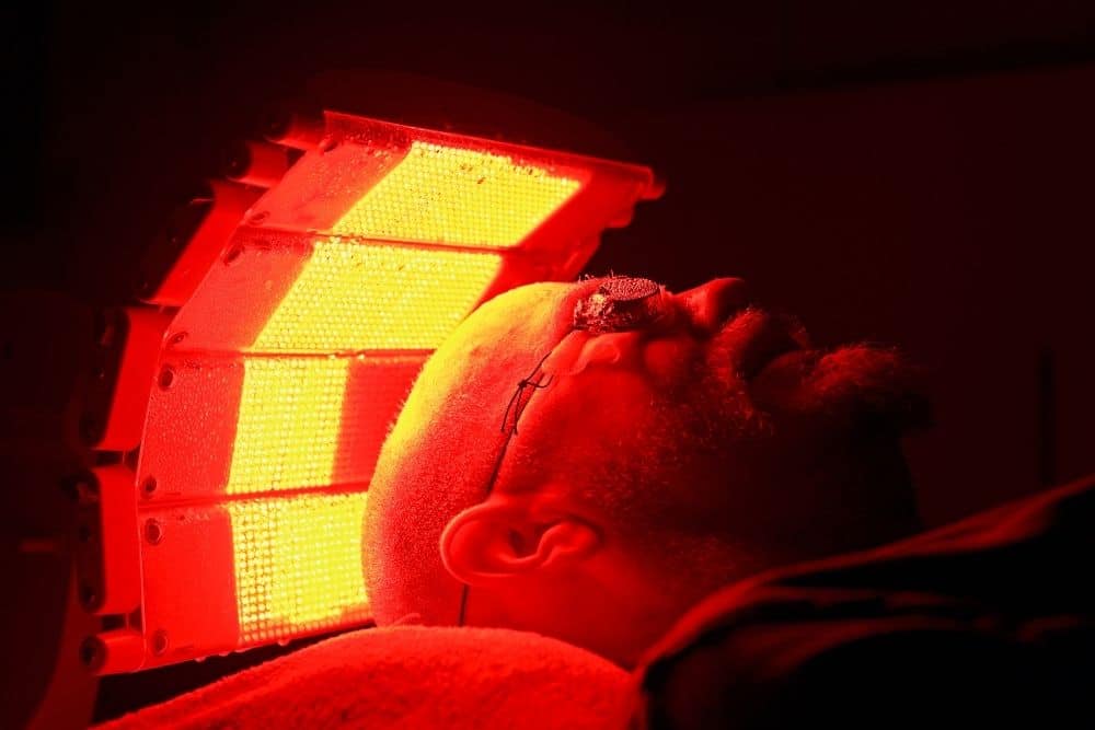 A person wearing eye protection lies under a red LED light therapy panel, illuminated by photodynamic therapy device in a dark room as part of a comprehensive strategy against extensive photodamage.
