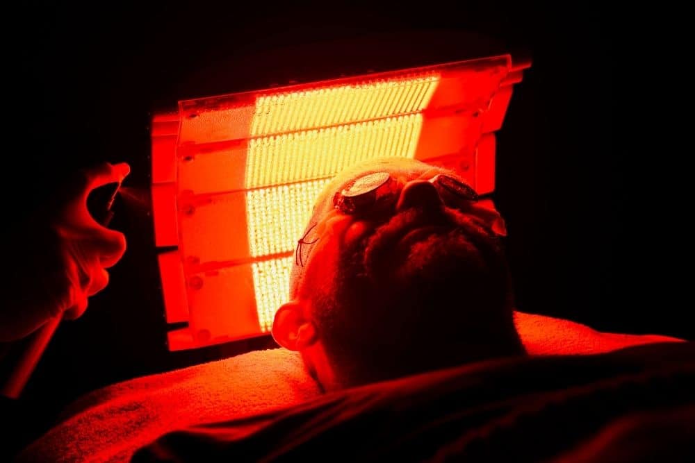 A person reclines under a red LED light therapy panel for PDT, wearing protective goggles in a dark room.