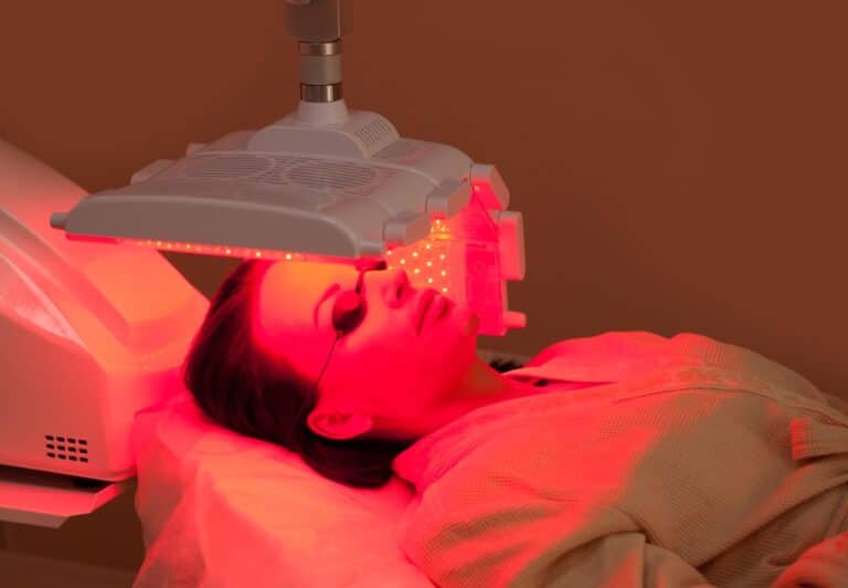 A person lies on a clinic bed receiving red light therapy on their face, wearing protective goggles under an LED light panel—a treatment often used in photodynamic therapy for skin cancer prevention.