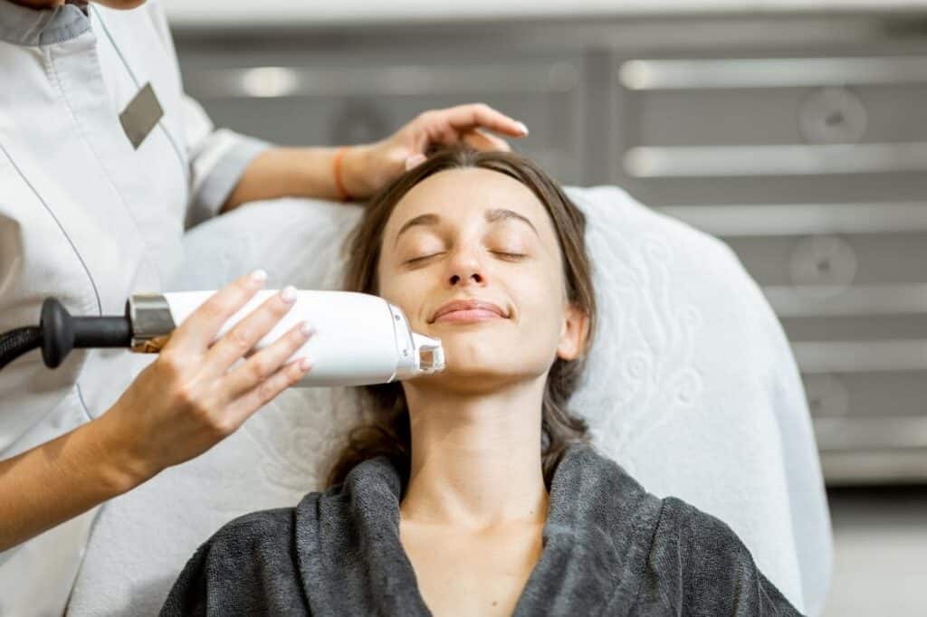 A woman is lying back with her eyes closed while a professional uses advanced fractional laser technology on her face in a clinical setting.