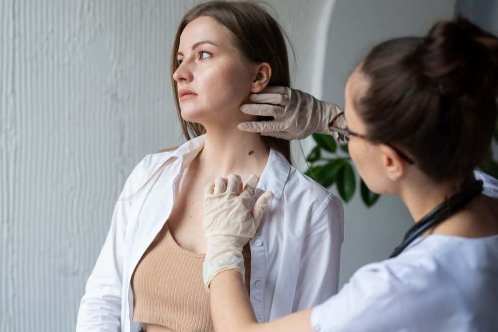 A female doctor wearing gloves examines a mole on a woman's neck and upper chest in a clinical setting, highlighting the importance of skin cancer prevention as part of a comprehensive strategy against extensive photodamage.