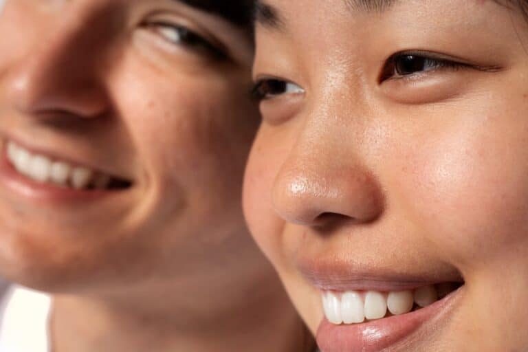Close-up of two people smiling, with focus on the face of the person on the right and the person on the left slightly out of focus in the background, beautifully highlighting how skin—the first organ of the body—glows with joy and connection.