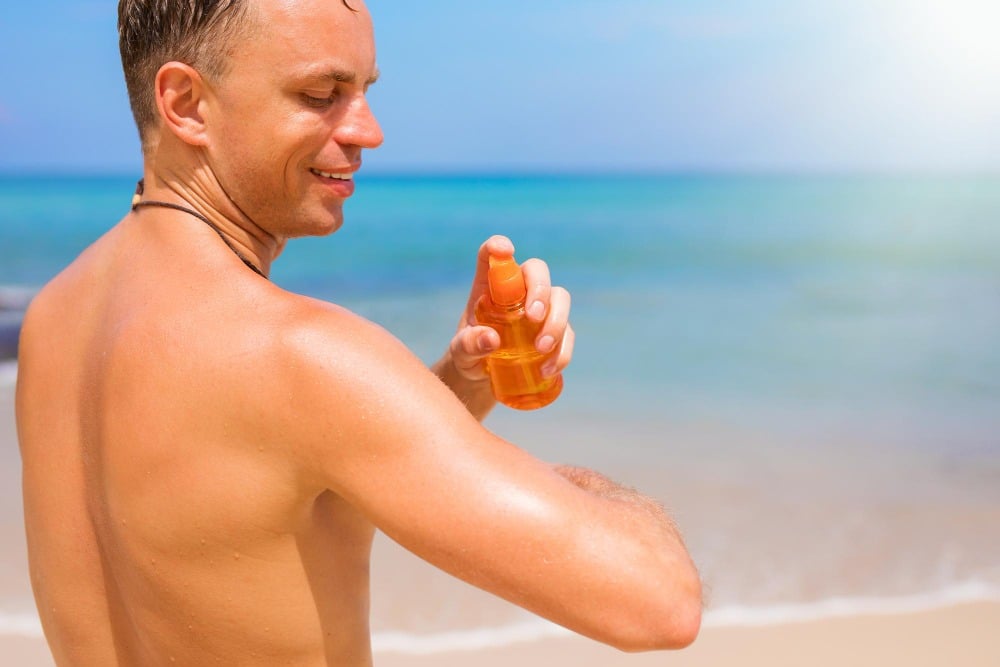 A man applies sunscreen spray to his shoulder while standing on a sandy beach with ocean waves in the background, reminding us how the history of skin and its evolution as our first organ has always provided vital protection against the sun’s rays.
