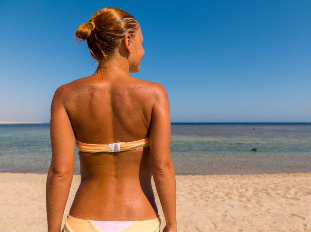 A woman in a bikini stands on a sandy beach, the resilient back skin of a woman facing the ocean with her back to the camera under a clear, blue sky.