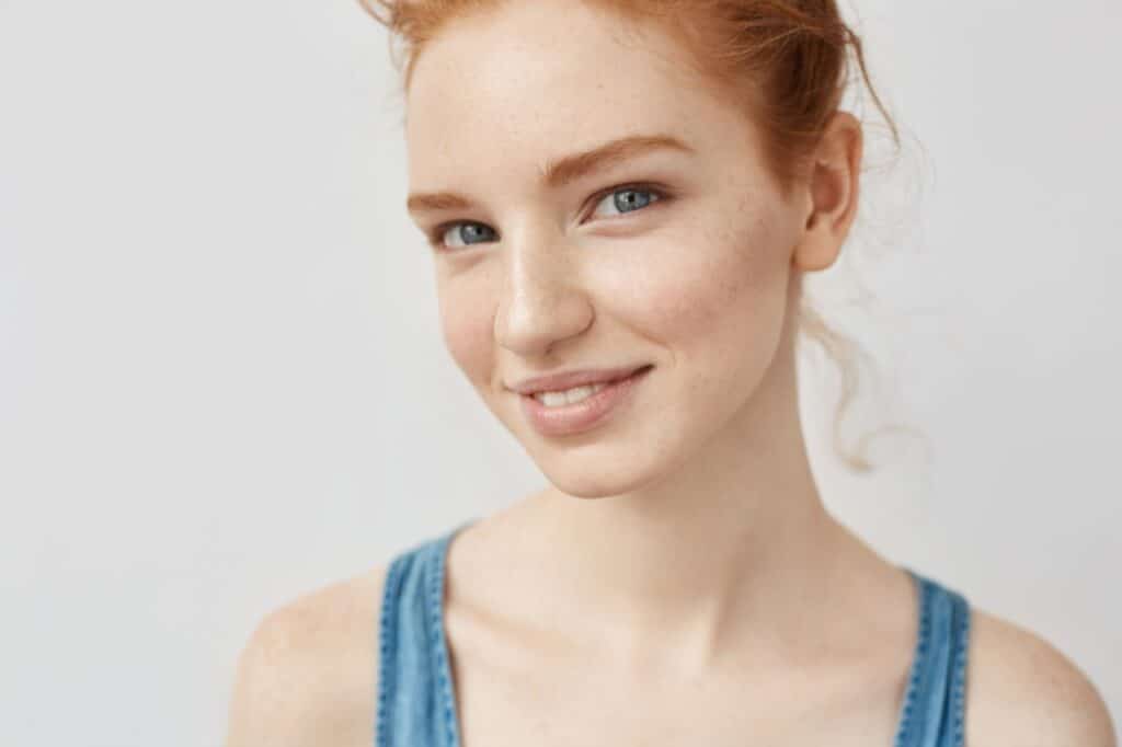 Young woman with red hair and fair skin, a classic feature associated with increased sun UV risk, wearing a blue sleeveless top and smiling slightly, posed against a plain white background.