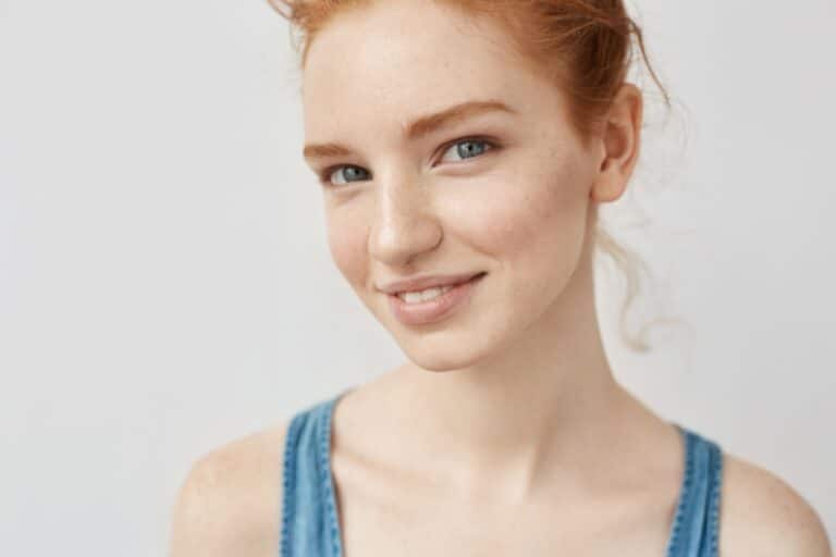 Young woman with red hair and fair skin, a classic feature associated with increased sun UV risk, wearing a blue sleeveless top and smiling slightly, posed against a plain white background.